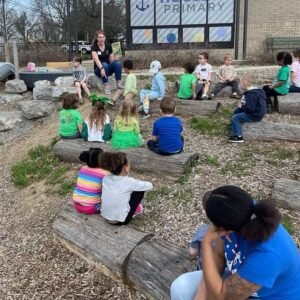 Students seated on logs in outdoor classroom, looking at educator reading "Animals in Camouflage" book.