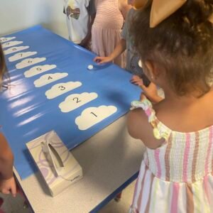 Blue mat on a table has clouds lined up with the numbers 1-10 printed on them. One student holds a small fan to blow a cotton ball.