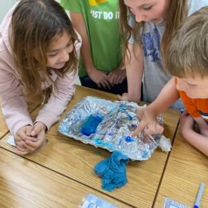 Students gathered around a foil model with blue water pooled. Students are looking and pointing at the places where water gathered.
