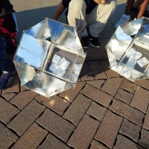 Two foldable solar ovens on a sidewalk. Students sit around ovens and look inside.